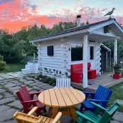 Sauna mit Sitzecke Weißes Saunahaus mit roter Gartenbank und bunten Adirondack-Stühlen im Garten, Abendhimmel in Rosa und Blau