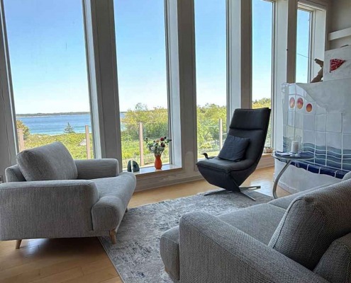 Salon lumineux avec de grandes fenêtres donnant sur la mer, aménagé avec des fauteuils modernes et un sol en bois clair.