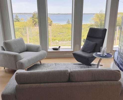 Salon moderne avec fenêtres panoramiques donnant sur l’océan, comprenant des fauteuils et une table d’appoint près d’un poêle en faïence blanc.