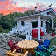 Saunaplace in the Backyard White Sauna cabin with red garden bench and colorful Adirondack chairs in a backyard, pink and blue evening sky above.