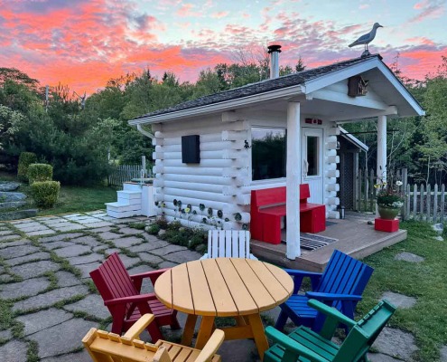 Saunaplace in the Backyard White Sauna cabin with red garden bench and colorful Adirondack chairs in a backyard, pink and blue evening sky above.