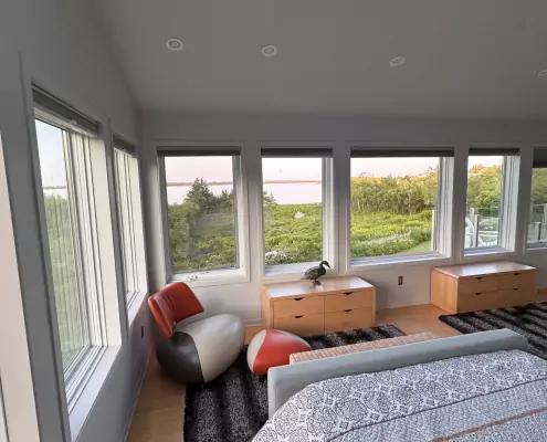Schlafzimmer mit Panoramafenstern Schlafzimmer mit Panoramafenstern und Blick auf grüne Natur und Wasser, gemütlicher Sessel mit Hocker am Fenster, moderne Einrichtung.