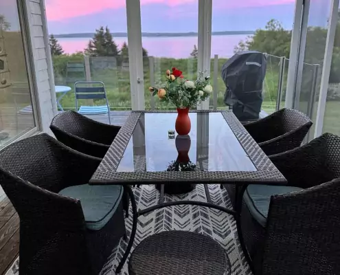 Abendstimmung auf der Terrasse Überdachte Terrasse mit Glastisch und Korbstühlen, Blick auf das Meer und rosa Abendhimmel.