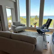 Gemütliches Wohnzimmer Gemütliches Wohnzimmer mit weitem Blick auf die Küste, stilvolle Einrichtung mit grauen Sesseln und einem schwarzen Relaxsessel vor einer großen Fensterfront.