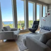 Heller Wohnbereich mit Blick aufs Meer Heller Wohnbereich mit bodentiefen Fenstern und Meerblick, ausgestattet mit modernen Sesseln und einem hellen Holzboden.