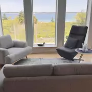 Wohnzimmer mit Panoramafenstern Modern eingerichtetes Wohnzimmer mit Panoramafenstern und Blick auf das Meer, Sessel und Beistelltisch neben einem weißen Kachelofen.